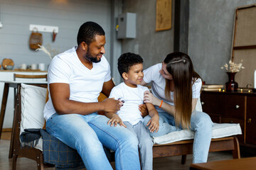Loving black family relaxing at sofa in the morning, happy mixed race parents laughing cuddling having fun with cute little kid child son playing enjoying moments together. High quality photo