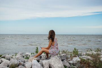 A beautiful young woman sits on a stone shore and looks at the ocean. Summer background.