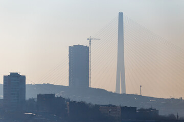 The silhouette of the Russian bridge across the Eastern Bosphorus is in dense fog. Russian bridge in Vladivostok against the background of residential buildings under construction.