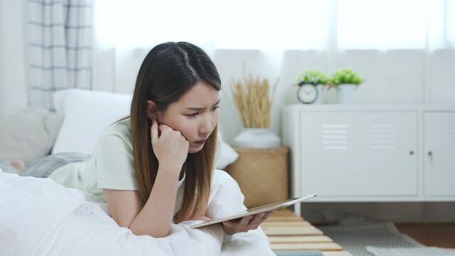 Thoughtful concerned Asian woman using tablet thinking something to solve the problems, imagining in mind to make the decision for her job in bedroom
