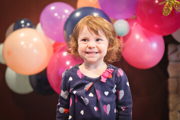 Close up Portrait of 2 Years Old Beautiful Toddler Girl with Big Blue Eyes and Brown Curly Hair. Happy Birthday girl, Blurry Colorfull Baloons Background