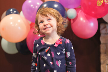 Close up Portrait of 2 Years Old Beautiful Toddler Girl with Big Blue Eyes and Brown Curly Hair. Happy Birthday girl, Blurry Colorfull Baloons Background