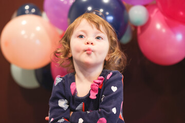 Close up Portrait of 2 Years Old Beautiful Toddler Girl with Big Blue Eyes and Brown Curly Hair. Happy Birthday girl, Blurry Colorfull Baloons Background