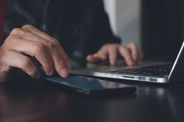 Man using mobile smartphone and touching on mobile phone screen on office table during working on laptop computer