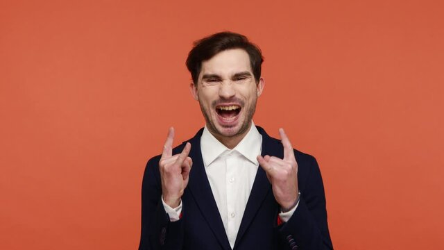 Enthusiastic Caucasian brunette guy in suit showing rock and roll sign gesture, rock symbol with fingers up, looking at camera, scream with excitement. Indoor studio shot isolated on orange background