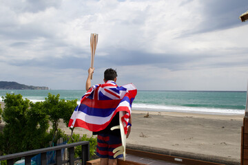 Man standing holding a flaming torch used in a large sporting event, with a surfboard and Hawaiian Flag looking at the Pacific Ocean in Chiba Japan where a big surfing competition is held.