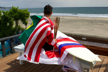 Man standing holding a flaming torch used in a large sporting event, with a surfboard and various Flags looking at the Pacific Ocean in Chiba Japan where a big surfing competition is held.
