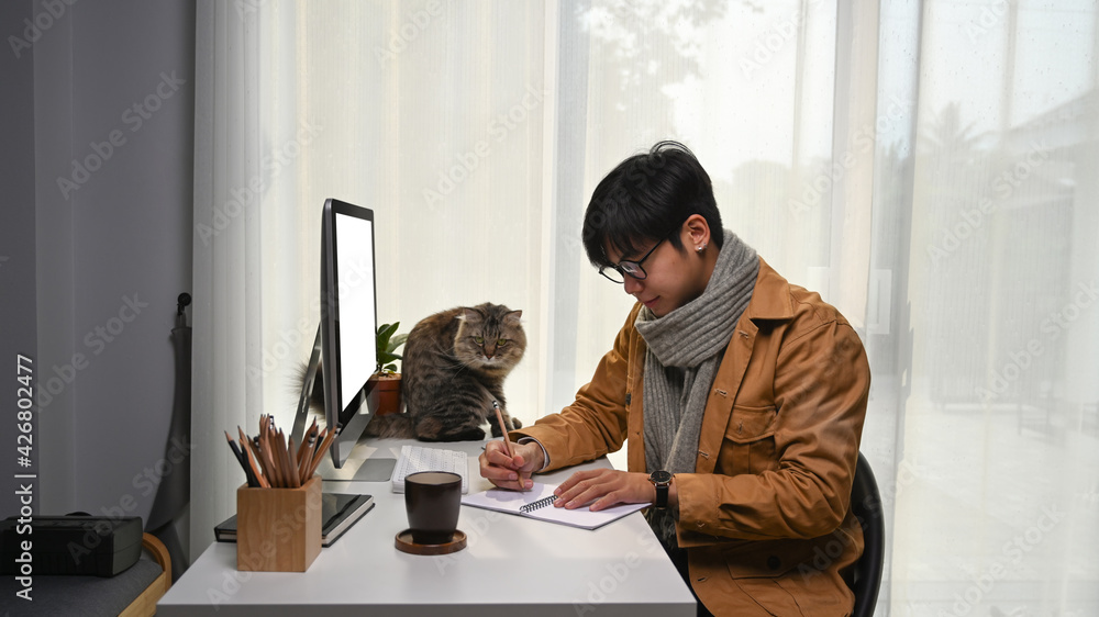 Poster young man sitting in front of computer with his cat and making note on notebook at home office.