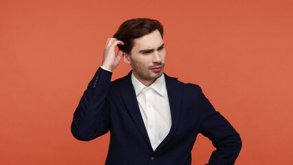 Frustrated young bearded businessman in suit scratching head thinks how to solve problem, has puzzled facial expression, has idea, raises finger up. Indoor studio shot isolated on orange background.