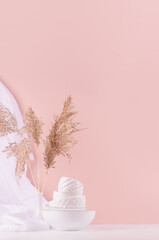 Delicate pastel pink interior with pile of white ceramic crockery, airy silk curtain and beige fluffy reeds on white wood table, vertical. Calm cozy wellness home decor.