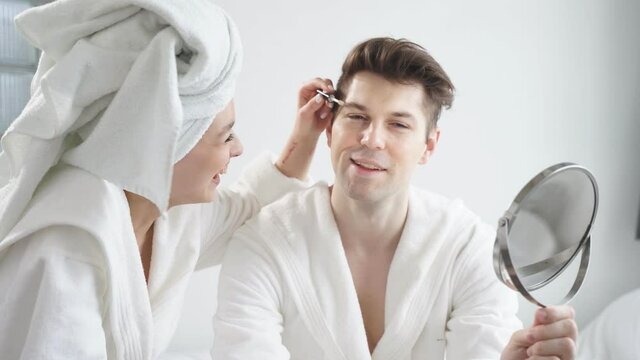 Portrait of funny couple doing make-up at home, woman doing make-up on face for man, they sit on bed after bathroom.
