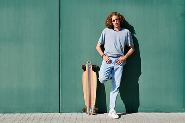 a young man with curly hair standing on a green wall on a sunny day with his skate or longboard