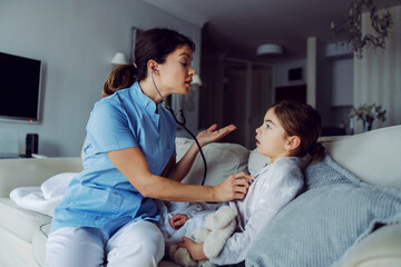 Doctor sitting on sofa next to girl and examining her lungs with stethoscope. Doctor at home service.