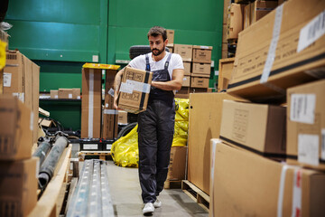 Hardworking tattooed bearded manual worker walking trough warehouse and carrying very heavy big box...