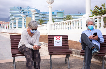 Fototapeta na wymiar Social distancing concept. Two senior people sitting outdoor on allowed bench, using mobile phone