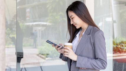 Young Asian woman wearing a gray suit is holding a phone and a paper coffee mug, Business women use smartphones to communicate by chatting or talking with business partners, Online media, VDO call.