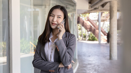 Fototapeta premium Young Asian female employee in a gray suit stands under the building of an office building talking on the phone, Talking using a mobile phone, Business communication, Smiling woman in a gray suit.