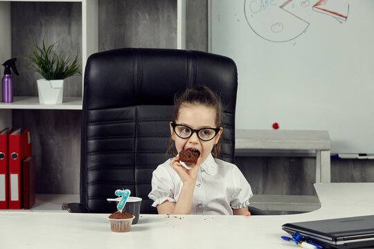 A little girl dressed as a businesswoman is sitting at an office desk eating a cupcake. The concept of business children. Children Are The Bosses. A board with a picture of a cake in the background.