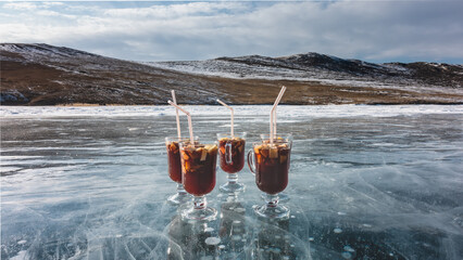 Glass mugs with mulled wine stand on a frozen lake. Red wine, sliced fruits, tubules. The ice is...