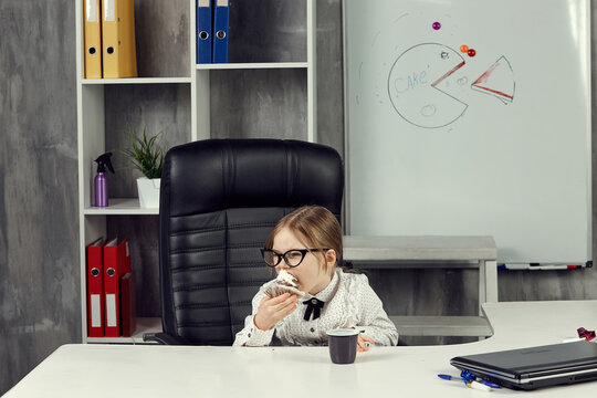 A little girl dressed as a businesswoman is sitting at an office desk eating a cupcake. The concept of business children. Children Are The Bosses. A board with a picture of a cake in the background.
