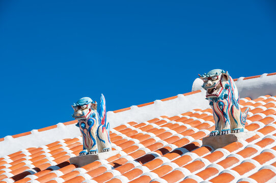 Two highly colored shisa statues, male and female, used as protection, on top of the red roof. Contrasting with the super blue sky in the background. Typical Okinawa scene.