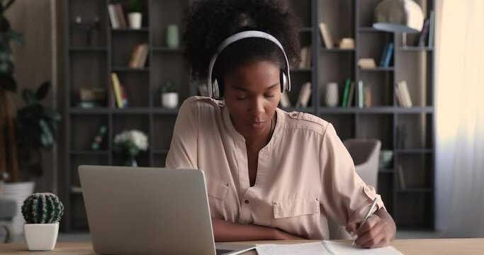 Happy attractive young biracial african woman in headphones involved in virtual educational event, holding web camera video call meeting with tutor, talking discussing distantly, writing notes.