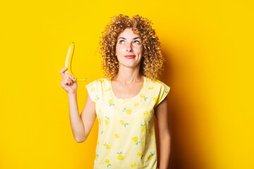 curly young woman holding a banana pensively looking upwards on a yellow background