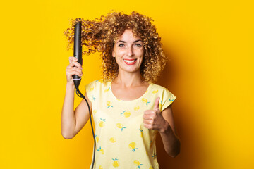 cheerful curly young woman straightens hair straightener on yellow background