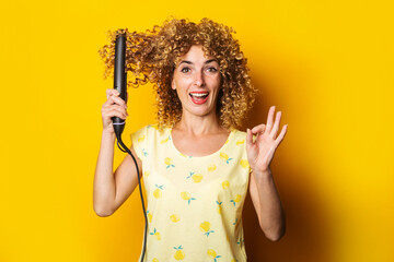 cheerful curly young woman straightens hair straightener on yellow background.
