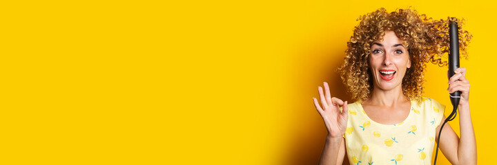 cheerful curly young woman straightens hair straightener on yellow background. Banner.