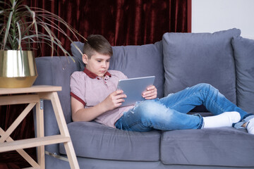 Young handsome teenager boy using tablet sitting on grey sofa at home