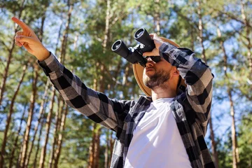 Tuinposter Man tourist in a hat and a plaid shirt looks through binoculars in the forest © Alex