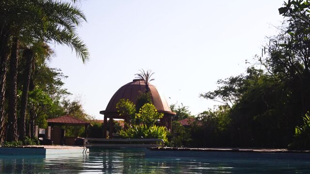 Establishing Shot Going From The Rippling Blue Water Of A Pool To The Domed Building In The Distance Surrounded By Trees With Birds Flying Overhead In Resort In Pushkar Rajasthan