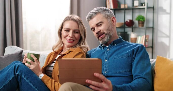 Close up of Caucasian beautiful middle-aged woman drinking coffee and speaking with bearded man typing on tablet device searching internet sitting on sofa in room, family couple, e-commerce concept