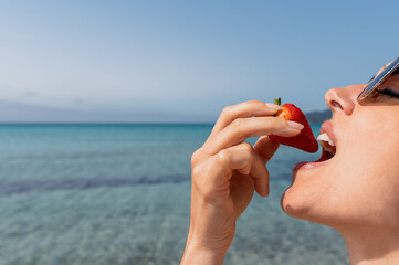Side view attractive young woman eating and biting a ripe strawberry with beautiful sea with clear waters background