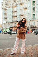 Fototapeta premium Vertical shot of a fashionable woman in sunglasses and stylish outfit hugging her small dog - york terrier, busy city street on a background.