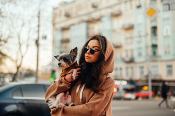 Urban shot of a fashionable woman in sunglasses and stylish hoodie hugging her small dog - york...