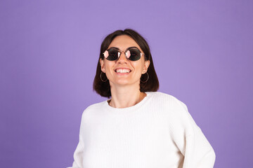 Young brunette in white casual sweater isolated on purple background wearing sunglasses happy smile positive moving