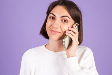 Young brunette in white casual sweater isolated on purple background having conversation with happy mood, talking positive on mobile phone, smiling
