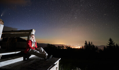 Female traveler in red jacket sitting on wooden steps under majestic colorful sky with stars....