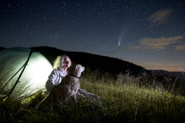 Young woman in the mountains observing beautiful starry night and a comet, sitting with her dog...