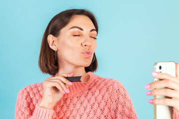 Young brunette in pink sweater isolated on blue background with makeup on face hold blush brush...
