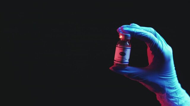 Pandemic immunization. Covid-19 prevention. Medical care. Vaccine dose vial in doctor hand in blue protective glove in red neon light isolated on black empty space background.