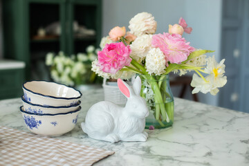 Bouquet of flowers in vase, Easter bunny and porcelain plates on table. Scandinavian style kitchen. Greeting card. Home interior with easter decor. Spring flowers in kitchen. Utensil on dining table