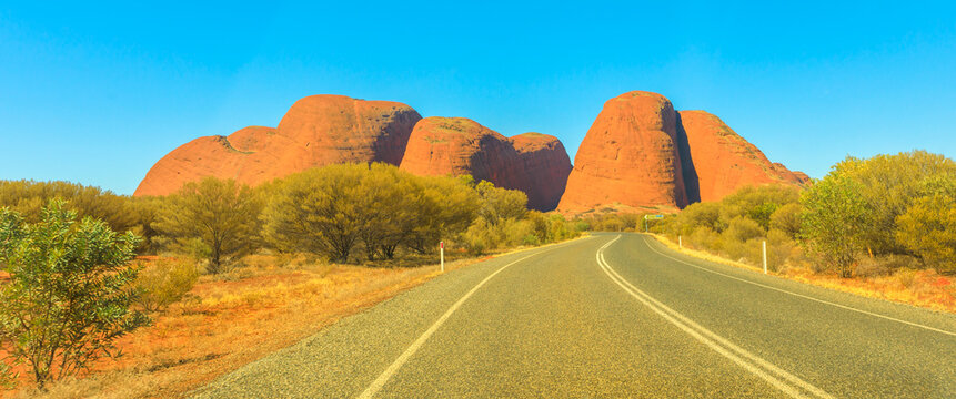 Uluru, Northern Territory, Australia - Aug 24, 2019: Panorama of road leading to domed rock formations in Uluru-Kata Tjuta National park at vibrant color of sunset. Aboriginal land and sacred places.