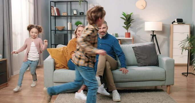 Cheerful Caucasian little cute children son and daughter running in living room playing having fun while mom and dad sitting on sofa together in positive mood, parents and kids, family concept