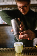 Bartender pours whiskey sour cocktail from metal shaker into glass through the sieve.