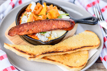 A serving of Shakshouka, a Maghrebi dish in a cast iron skillet, with eggs poached in a sauce of tomatoes, olive oil, peppers, onion and garlic. Served with s spicy sausage and slices of bread