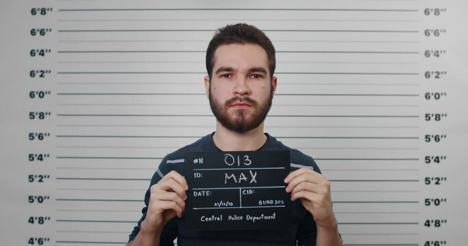 Portrait of young guy with mustaches and beard holding sign for photo in police department. Crop view of millennial man posing, raising head and looking to camera.Concept of criminal.