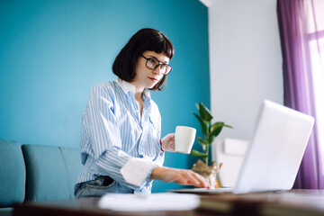 Beautiful  woman working on laptop computer while sitting at the living room, drinking coffee. Technology, freelance, shopping online, remote work, E learning concept. 